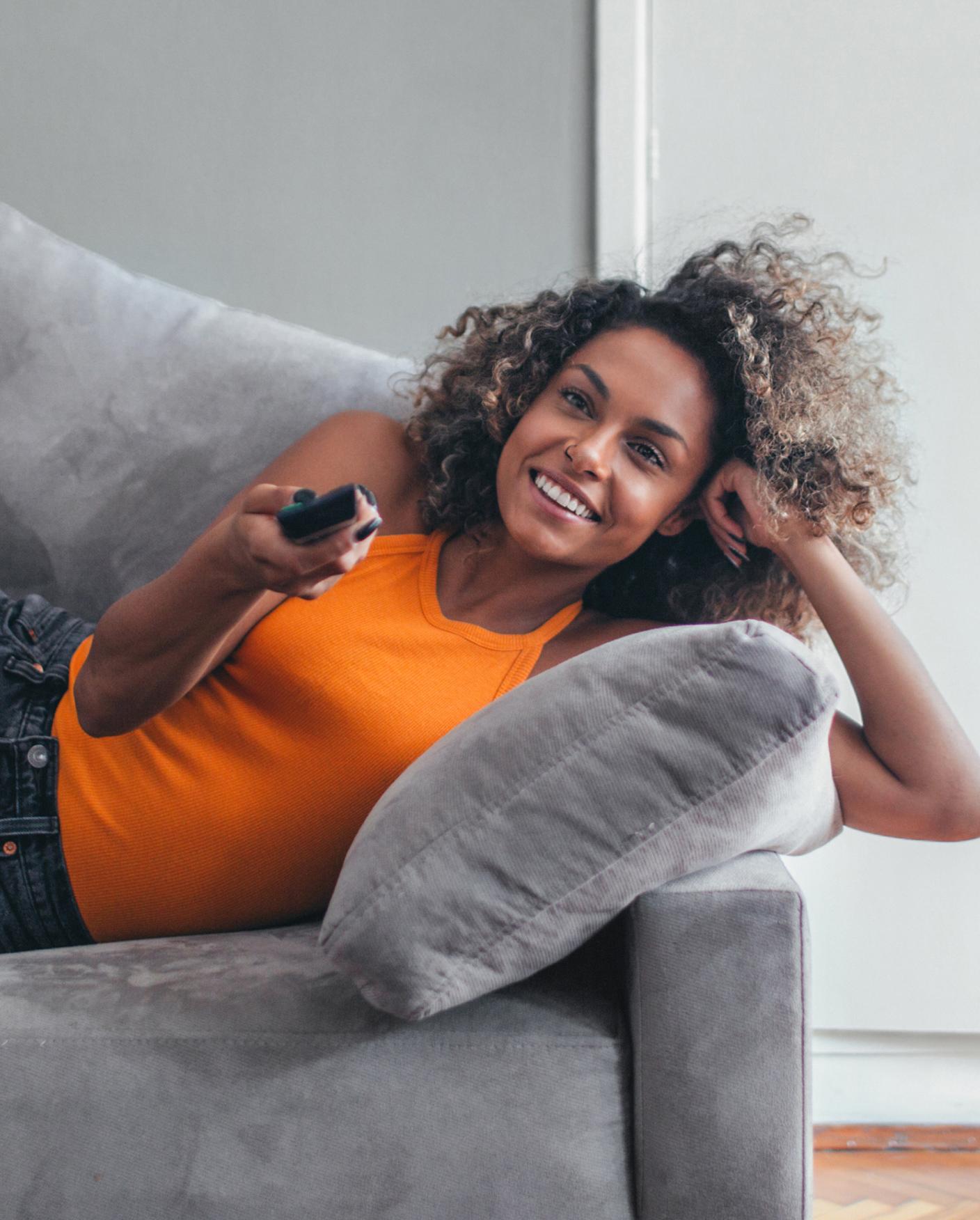 woman lays on grey couch holding remote control and changing channel on tv