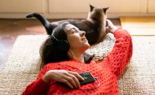 young girl lounging on living room floor, listening to headphones, holding phone and petting cat.