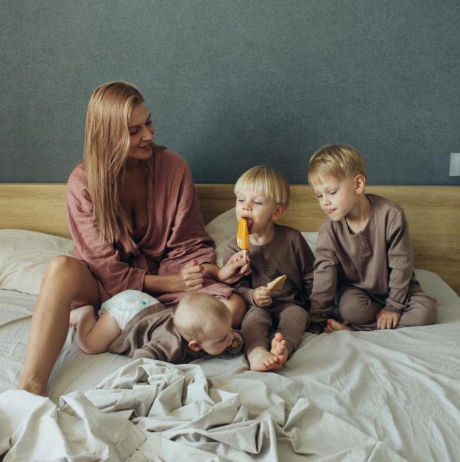 Adult and three young children sitting and playing together on a bed in a bedroom. One child is eating a popsicle, another is holding a snack, and a baby is lying on the bed in the foreground